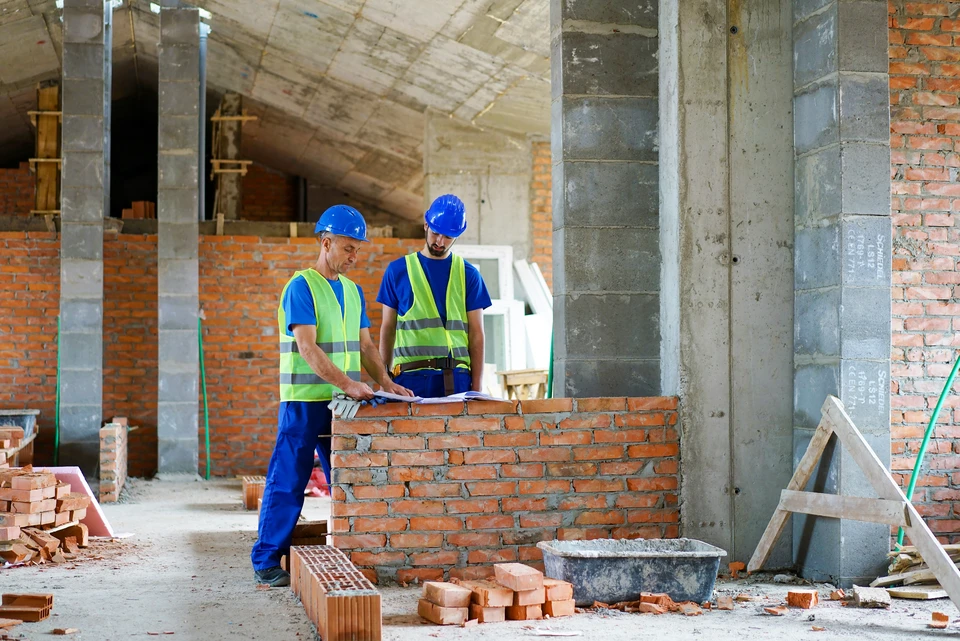 Chantier de construction intérieur avec ouvrier souriant en casque de sécurité blanc et combinaison bleue, portant gants orange, appuyé sur surface
